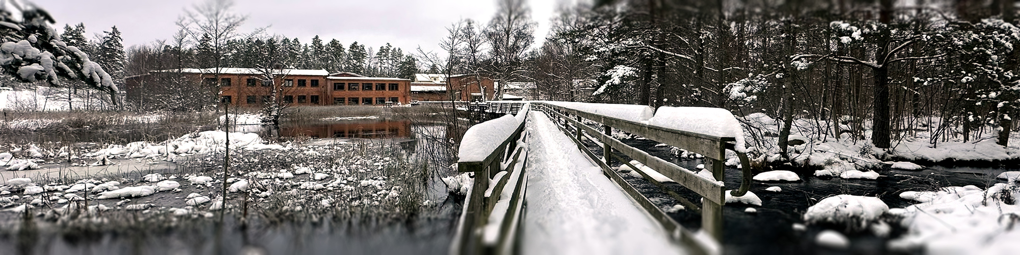 Snötäckt träbro över Alsterån i Fröseke under vintertid, med vattendrag, vass och en röd tegelbyggnad i bakgrunden.