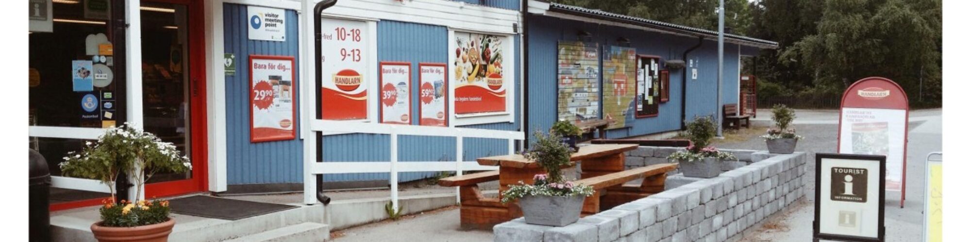 Lanthandel i en blå träbyggnad med skyltar och entré mot gatan. Utanför finns en liten sittplats med träbord och bänkar, planteringar i krukor samt informationsskyltar, vilket skapar en mötesplats i bymiljö.