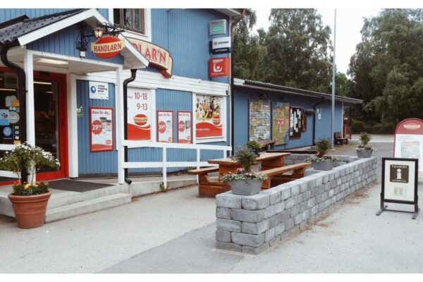 Lanthandel i en blå träbyggnad med skyltar och entré mot gatan. Utanför finns en liten sittplats med träbord och bänkar, planteringar i krukor samt informationsskyltar, vilket skapar en mötesplats i bymiljö.