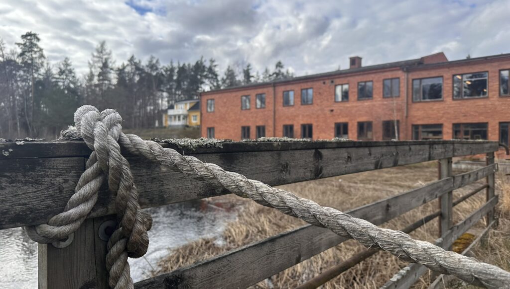 Rep knutet runt ett trästaket vid Alsterån i Fröseke, med en röd tegelbyggnad och vattendrag i bakgrunden.