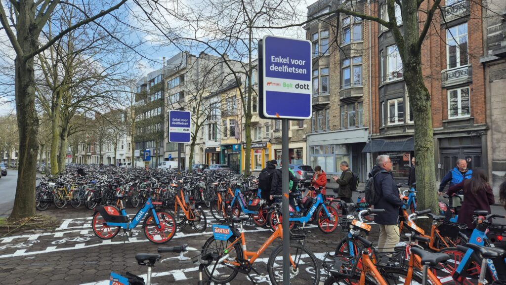 Cykelparkering i stadsmiljö med många parkerade cyklar och delade cyklar, samt personer som rör sig i området.