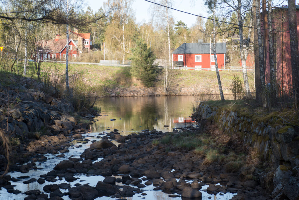 Vattendrag med stenar och lugnt vatten omgivet av träd och röda hus i en småortsmiljö i Fröseke.