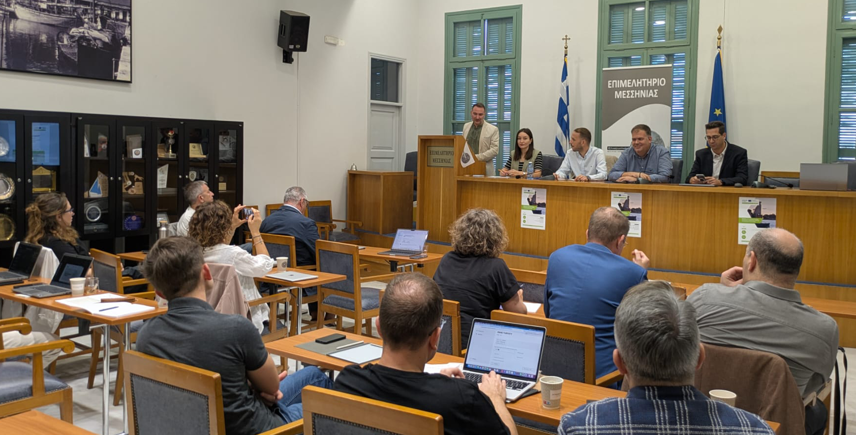 Deltagare från PARSMO-projektets internationella partnermöte i Kalamata, Grekland, sitter i en konferenssal och lyssnar på lokala representanter som välkomnar gruppen vid ett podium. Bakom talarna syns Greklands och EU:s flaggor.
