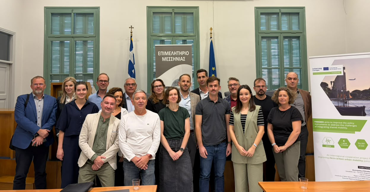 Gruppfoto från PARSMO-projektets internationella partnermöte i Kalamata, Grekland. Deltagare från sex länder står tillsammans i en konferenssal framför Greklands och EU:s flaggor, med en informationsrollup om projektet till höger.